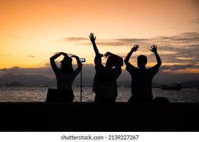 Silhouette Of Youngsters Students Boy And Girl Chat And Have Fun And Take Selfie Of Mobile Phone Camera During Sunset In West Kowloon Waterfront Promenade, Hong Kong In Evening. Back Light Shot