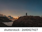 A silhouetted couple stoop atop a rocky hilltop by the sea at dusk gazing out at the moon and cloud glowing under the first lights of the day
