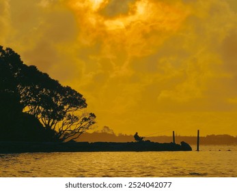 A silhouetted fisherman fishing and relaxing in the evening on a chair beside the estuary waters at sunset with trees behind him - Powered by Shutterstock