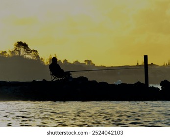 A silhouetted fisherman fishing and relaxing in the evening on a chair beside the estuary waters at sunset - Powered by Shutterstock