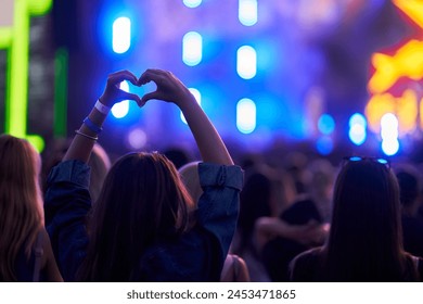 Silhouetted hands form heart shape at vibrant music fest. Audience enjoys live concert, colorful stage lights in background. Festive atmosphere, fan gestures love, joins crowded outdoor event.