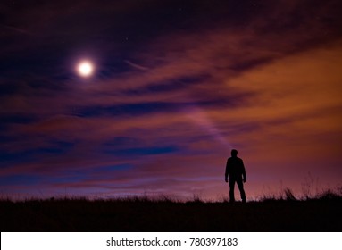 Silhouetted Person Stands Atop A Grassy Hill With A Light Beam Looking Up At The Milky Way, Moon, And Colored Clouds At Sunrise.