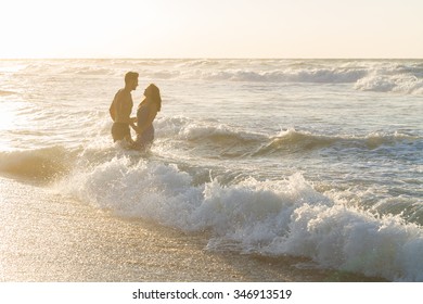 Silhouetted young couple at the beach on a  hazy summer day at dusk, in a blue dress and shorts, enjoying  splashing in the ocean water, getting wet, teasing and kissing one another. - Powered by Shutterstock