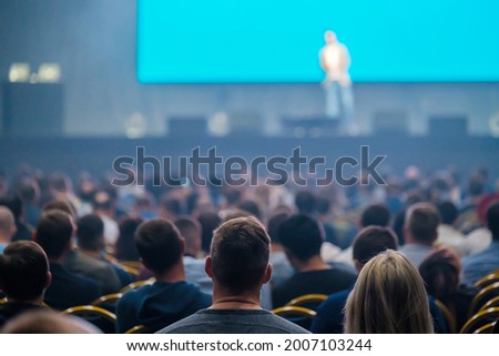Silhouettes of anonymous people sitting in dark auditorium and listening to speaker talking from stage during business seminar
