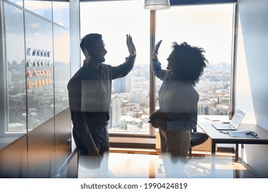 Silhouettes of cheerful successful business partners indian businessman and African American businesswoman colleagues giving high five celebrating business triumph in office at panoramic window.