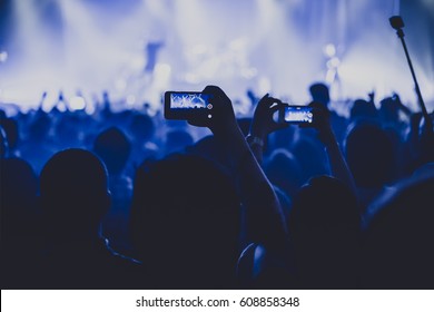 Silhouettes Of Concert Crowd In Front Of Bright Stage Lights. Dark Background, Smoke, Concert  Spotlights. Group Of People Holding Hands With Mobile Phones At A Concert 