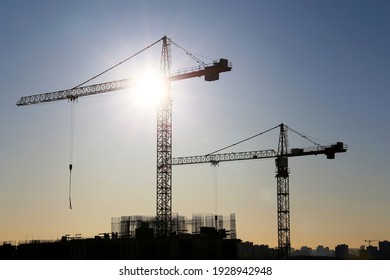 Silhouettes of construction cranes and unfinished residential buildings on blue sky and sunshine background. Housing construction, apartment block in city