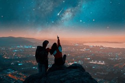 Silhouettes D'un Couple Assis Au Sommet De La Montagne Regardant Et Pointant Vers L'étoile Filante Et La Voie Lactée Au-dessus Des Lumières De La Ville à L'horizon	