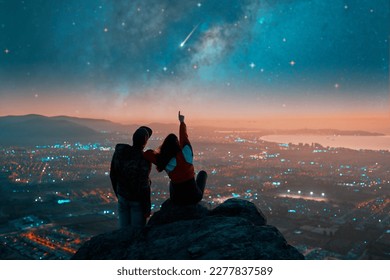 silhouettes of a couple sitting on the top of the mountain looking and pointing out at shooting star and milky way over the city lights on the horizon	