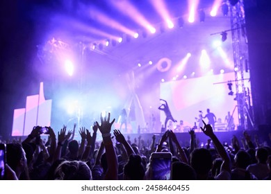 Silhouettes of excited crowd at music fest, hands raised with lit stage in background. Band performs at night, colorful lights illuminate event. Fans enjoy live concert, moments on smartphones. - Powered by Shutterstock