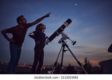 Silhouettes of father, daughter and astronomical telescope under starry skies.