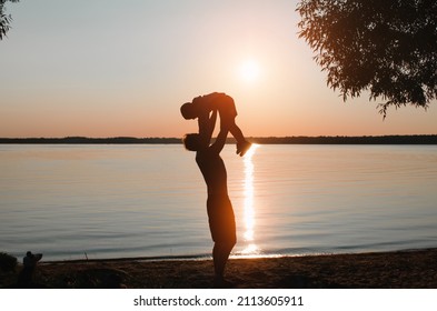 Silhouettes Of Father And Son Having Fun On Shore Of Lake At Sunset, Outdoors. Side View Of Father Holding Child In Arms, Throwing Up Air Against Sky. Fatherhood, Childhood, Family Vacation.