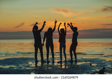 Silhouettes of four diverse young friends dancing with beer bottles in raised hands by sea at orange sunset during beach party, backlit back view - Powered by Shutterstock