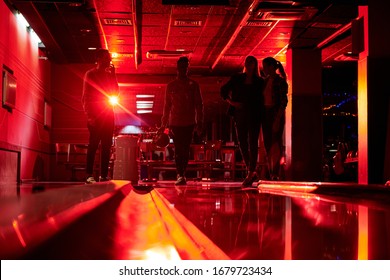 Silhouettes Of Four Young People Standing By Bowling Alley In Dark Room While One Of Guys Going To Throw Ball