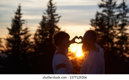Silhouettes of happy couple in love in the evening. Bearded man and smiling woman gesturing heart shaped hands, looking at each. Beautiful sunset, cloudy sky and forest on blurred background