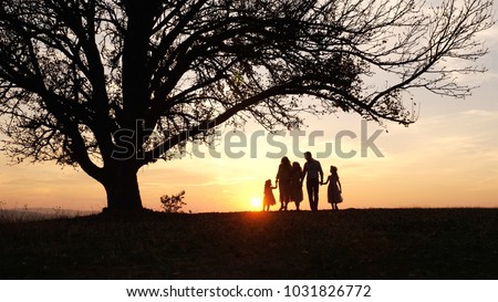 Silhouettes of happy family walking in the meadow near a big tree during sunset. Family spent time together. Enjoing each other