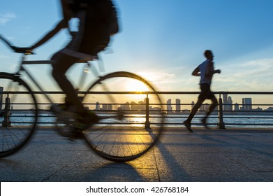 Silhouettes Of Jogger Running And Bicyclist Cycling At Sunset In Front Of The City Skyline 