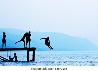 Silhouettes Of Kids Who Jump Off Dock On The Lake.Blue Toned.