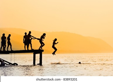 Silhouettes Of Kids Who Jump Off Dock On The Lake At Sunset.