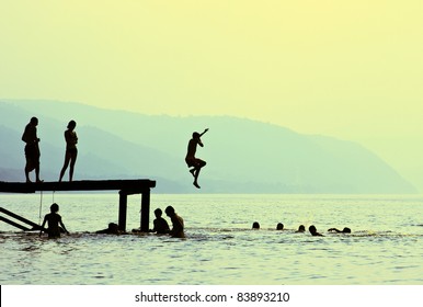 Silhouettes Of Kids Who Jump Off Dock On The Lake At Sunset.