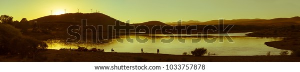 Silhouettes of people in lake or marsh landscape,\
at sunset after a hill with wind turbines, collect belongings in\
cars at the end of a day of summer excursion in the water. Photo\
treatment in\
monochr