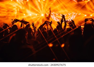 Silhouettes of people rising their hands and dancing on on open air music festival at night while performer on stage is making a good atmosphere. There are orange stage lights. Nightlife, concerts. - Powered by Shutterstock