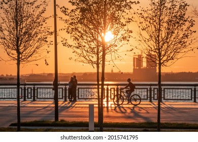 Silhouettes of people walking along the embankment against the background of the setting sun and glare on the water. Sunset on the city promenade.