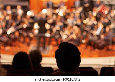 Silhouettes Of Spectators In The Foreground And A Concert Of Classical Music Performed By A Symphony Orchestra In A Hall Blurred. Well-lit Background, Scene With Many Lights.