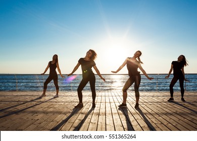 Silhouettes of sportive girls dancing zumba near sea at sunrise.