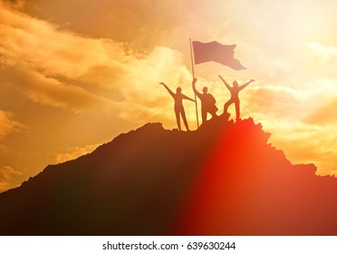 Silhouettes Of Three People Holding A National Flag Of Victory On A Man On Top Of A Mountain, Hands Up. Conceptual Design. Against The Dramatic Sky With Clouds At Sunset. Business Success.