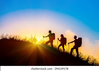 Silhouettes of three people walking with backpacks and other hiking gear up toward top of wild grass mountain mother father daughter bright luminous sunrise sky background 