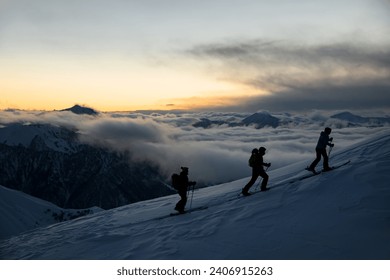 Silhouettes of three skiers with poles going up the slope against the background of mountain peaks covered with fog at sunset