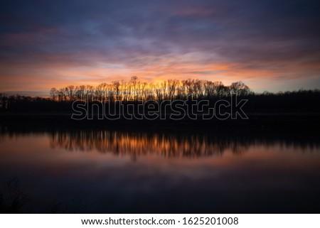 The silhouettes of trees under the cloudy sunset sky reflected in the lake below