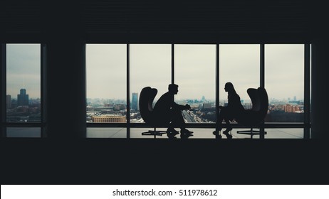 Silhouettes Of Two Business People Man And Woman Sitting On Armchairs In Office Interior, Man Showing Information On Screen Of His Laptop To His Colleague, Huge Windows With Winter Cityscape Outside