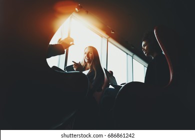 Silhouettes Of Two Business Women In Dark Office Settings Sitting On Armchairs With Their Gadgets And Having Lively Discussion With Their Third Colleague During Work Meeting Near Window