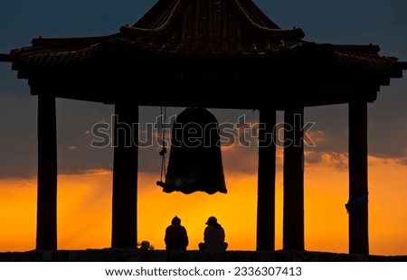 The silhouettes of two people sitting under a Buddhist prayer bell in the backlight of the orange evening sky in the Gobi desert, Mongolia