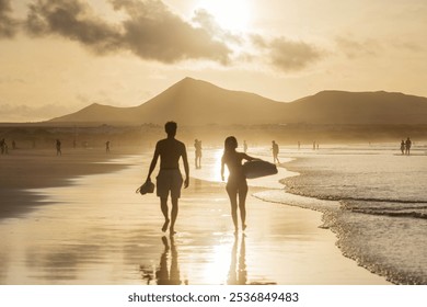 Silhouettes of two surfers walking along Famara Beach at sunset in Lanzarote, with their shadows stretching across the sand. - Powered by Shutterstock