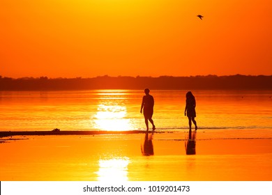 Silhouettes of two wooman (mothers and daughters) walking through the shallows of a lake against the background of a red-orange sunset - Powered by Shutterstock