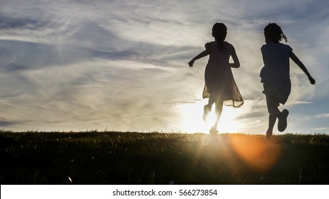 Silhouettes Of Two Young Children Running Towards The Sunset Over A Hilltop