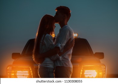 Silhouettes of two young people in the light of burning car headlights in the background. The guy hugs and kisses the girl on the forehead, saying goodbye to her before leaving for against his car.