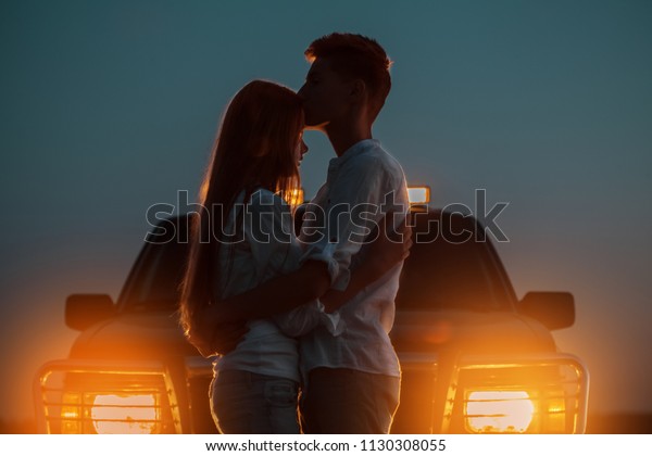 Silhouettes of two young people in the light of\
burning car headlights in the background. The guy hugs and kisses\
the girl on the forehead, saying goodbye to her before leaving for\
against his car.