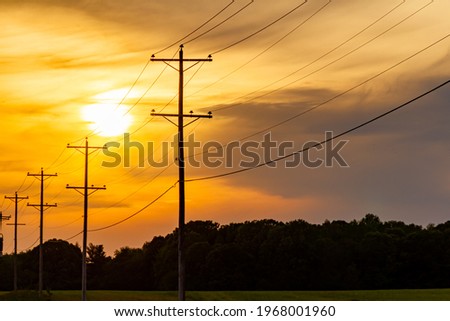 Silhouettes of utility poles carrying high voltage electricity alongside a road at a rural area. Sunset, sunrise image with cloudy orange red sky and depth of field with nobody in the frame.
