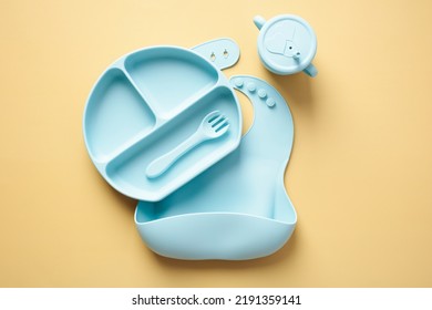 Silicone Baby Feeding Supplies Set. Top View Silicone Bib, Plate With Fork, Sippy Cup On Pastel Yellow Background. First Feeding, Baby Led Weaning Concept.