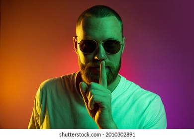 Sillent  quiet  Portrait handsome young man in sunglasses and finger to lips isolated over gradient orange pink background in neon lights  Concept human emotions  facial expression  mood 