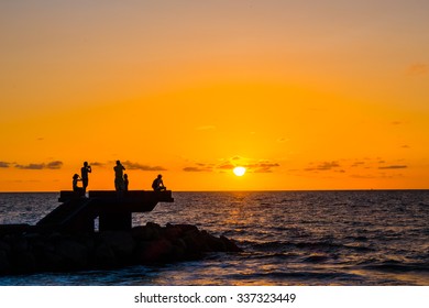 Sillhouette Of Group Of Friends On A Lookout Point On A Rock In The Beach During Sunset