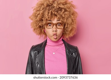 Silly funny woman with curly hair puckers lips makes grimace wears spectacles turtleneck and leather jacket poses against pink background imitates fish being childish does facial expressions