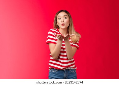 Silly glamour tender asian blond modern girl send you sweet kisses. Joyful urban woman wear striped t-shirt hold palms near fold lips blow kiss air mwah, stand red background glamour