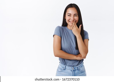 Silly Hideous Female Asian College Student Making Prank And Giggle In Corner As Watching Someone Got Tricked, Look Sle Left, Cover Smiling Mouth With Hand And Chuckling Secretly, White Background