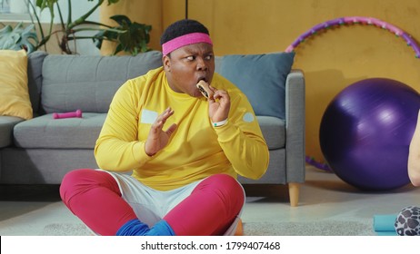 Silly Overweight Black Man Sitting In Lotus Pose And Eating Cookie Without Permission During Meditation Session With Personal Coach. Concept Of Fun, Obesity, Junk Food.