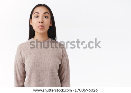 Silly and playful cute asian woman in winter sweater acting childish and funny, showing tongue, pouting and look camera tenderly, fool around, making goofy expressions over white background